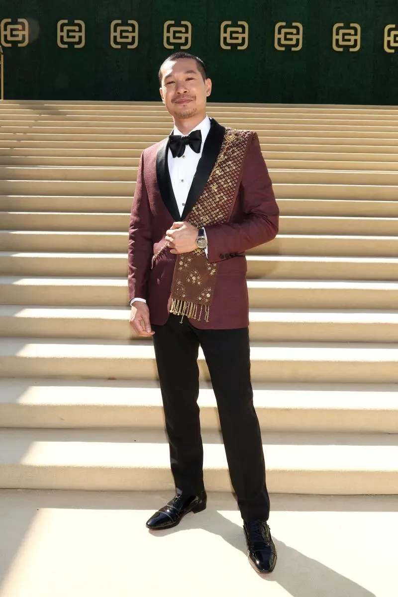 مخرج يرتدي بدلة أنيقة باللون البني الداكن مع وشاح مزخرف، يقف على درجات سلم أمام خلفية خضراء تحمل شعار علامة تجارية.