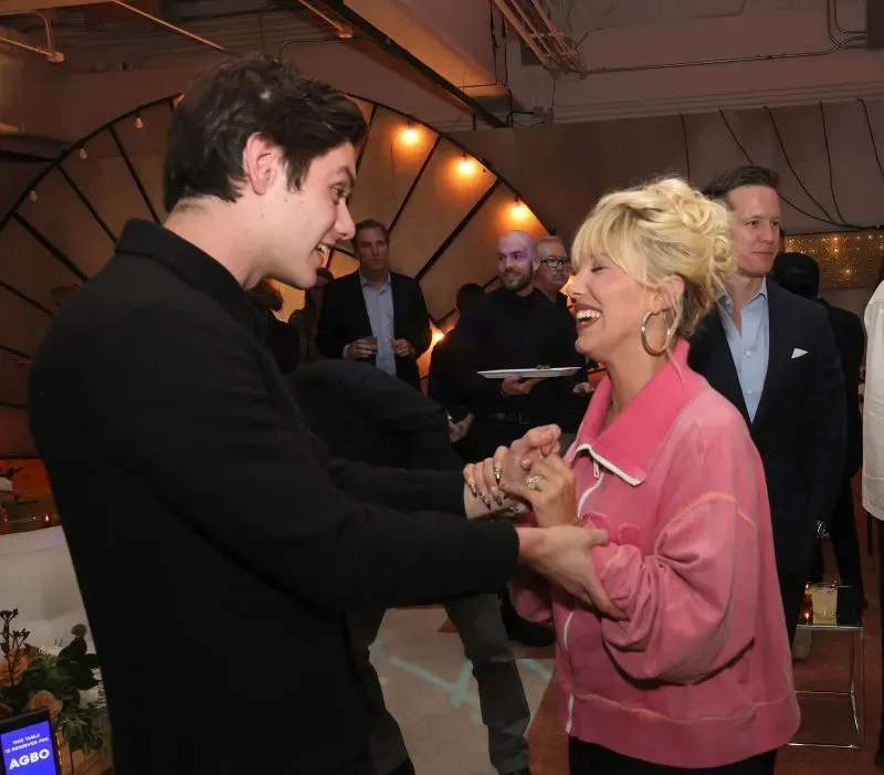 ميلي بوبي براون تتفاعل مع الحضور في حدث، مرتدية سترة وردية وتبتسم، مما يعكس أسلوبها المستوحى من التسعينيات.