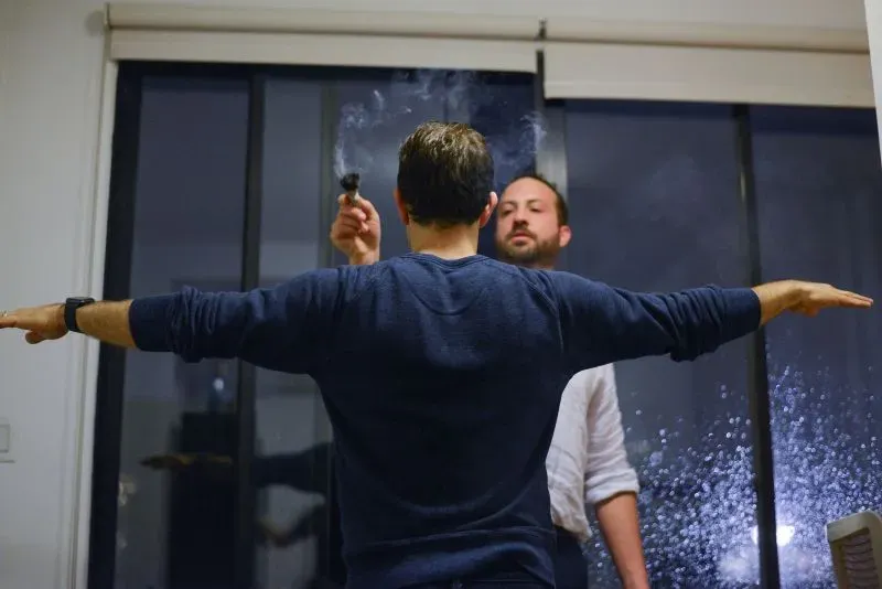 رجل يقف مع ذراعيه ممدودتين بينما يقوم آخر بإشعال بخور، في إطار نشاط جماعي يهدف لتعزيز التواصل العاطفي بين الرجال.