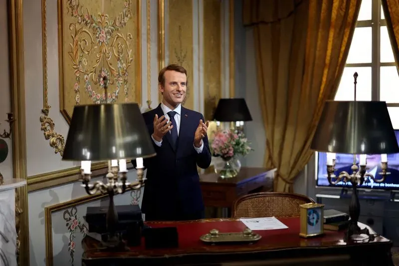 تمثال للرئيس الفرنسي إيمانويل ماكرون مبتسمًا ويصفق، موضوع في مكتب مزين بألوان دافئة، في سياق احتجاج على سياسته البيئية وعلاقاته التجارية مع روسيا.