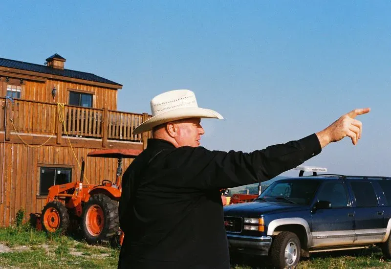 رجل يرتدي قبعة رعاة البقر يشير بيده نحو الأفق، خلفه منزل خشبي ومعدات زراعية، يعكس روح الاستقلال في ألبرتا.