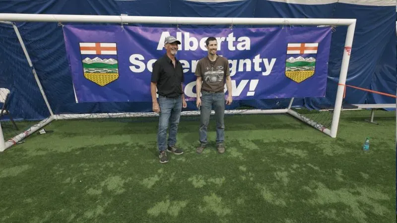 رجلان يقفان أمام لافتة كبيرة تحمل عبارة "استقلال ألبرتا الآن!" في منشأة رياضية، مع أجواء تجمع مؤيدين للاستقلال.