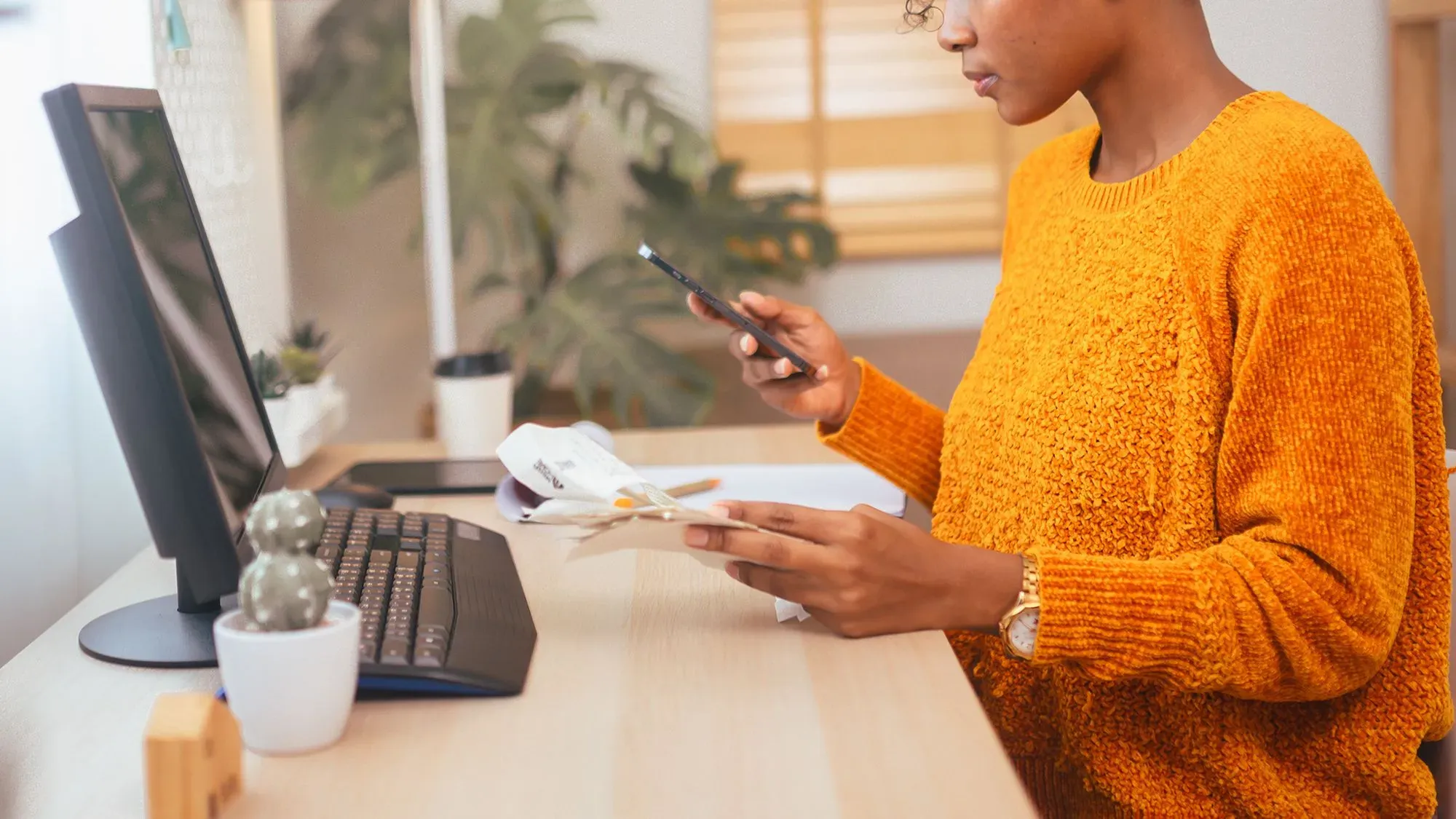 امرأة ترتدي سترة برتقالية تجلس أمام جهاز كمبيوتر، تحمل هاتفًا في يدها وتراجع مستندات مالية، في بيئة مكتبية مريحة.