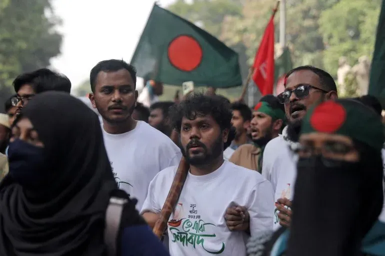 محتجون في شوارع دكا يحملون أعلام بنغلاديش، يرتدون قمصانًا مكتوب عليها شعارات سياسية، مع تعبيرات حماسية تعكس روح الانتفاضة.