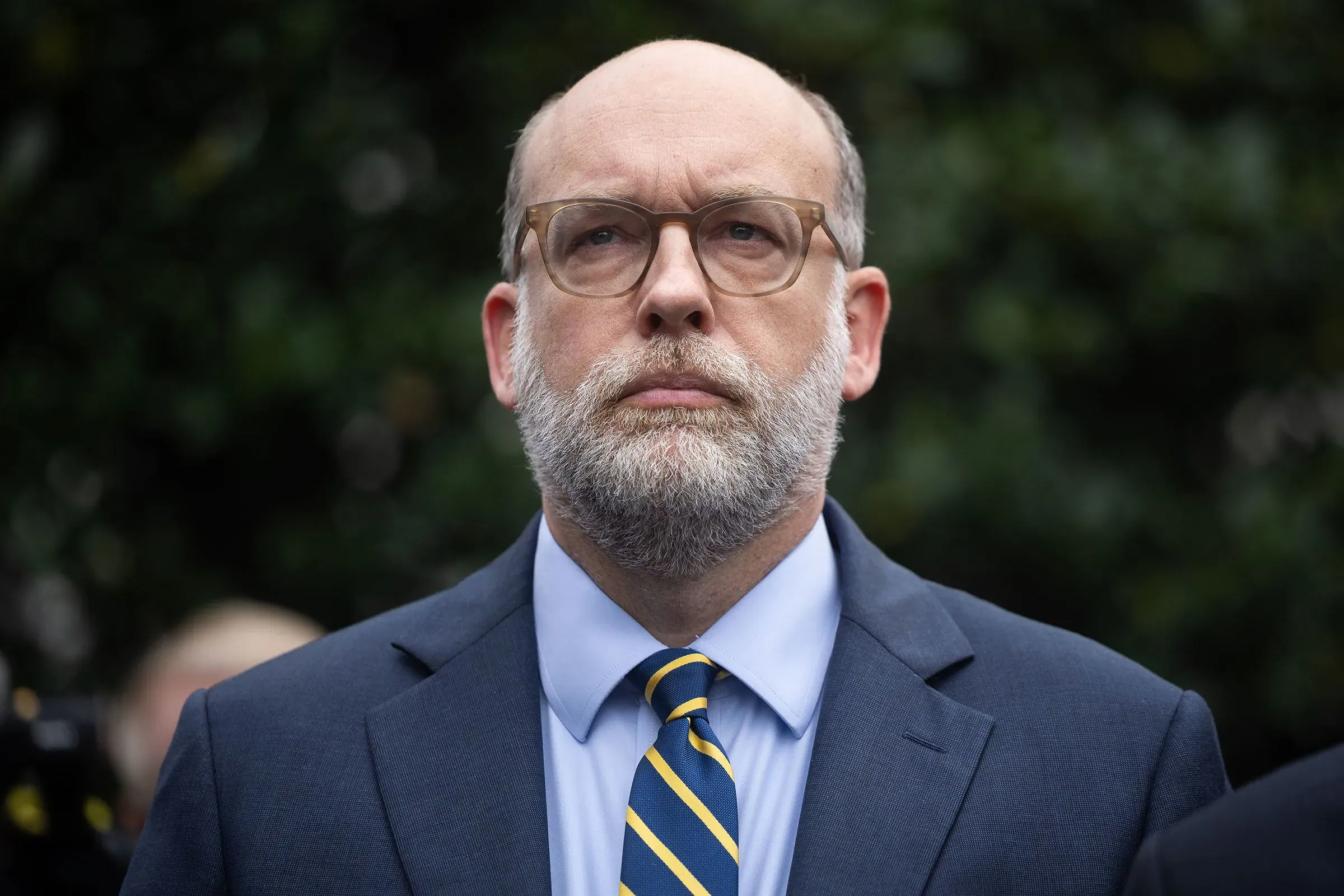 رجل ذو لحية يرتدي بدلة رسمية ونظارات، يظهر بملامح جدية، في خلفية طبيعية، في سياق مرتبط بالتحقيقات الجنائية.