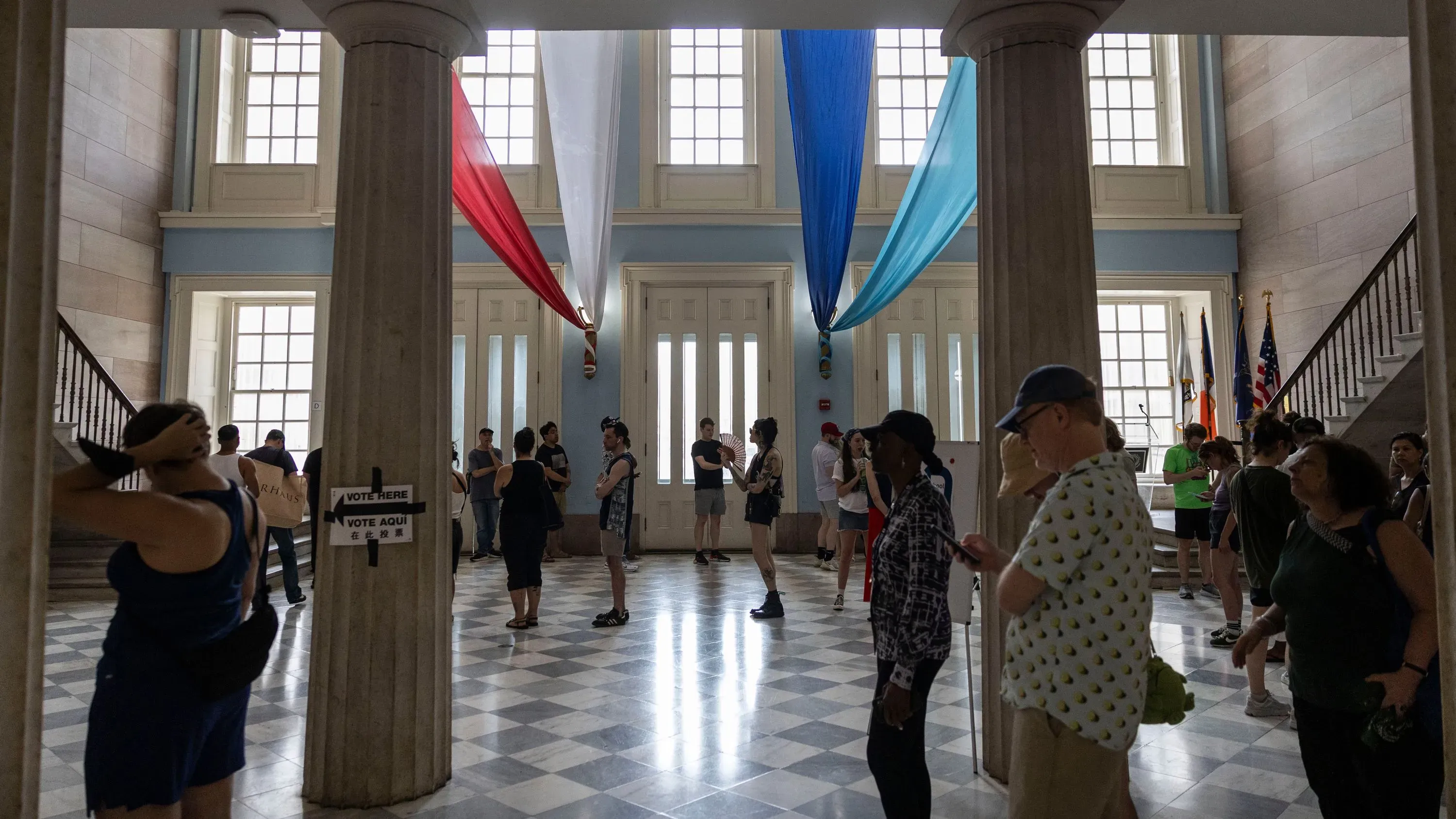 مواطنون يقفون في طابور داخل مبنى حكومي للتصويت، مع أعلام ملونة تتدلى من السقف، يعكس تنوع الآراء السياسية في الانتخابات.