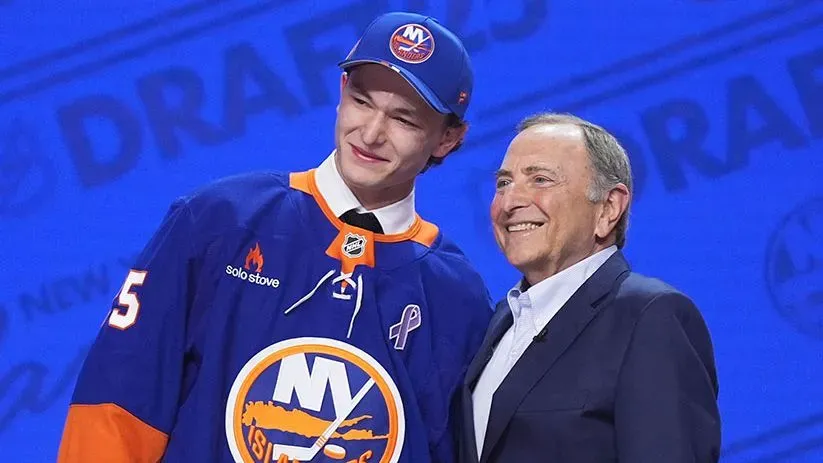 ماثيو شايفر، لاعب هوكي كندي، يقف مبتسمًا بجوار مفوض NHL غاري بيتمان بعد اختياره كأول اختيار في مسودة 2025، مرتديًا سترة آيلاندرز مع شريط لسرطان الثدي.
