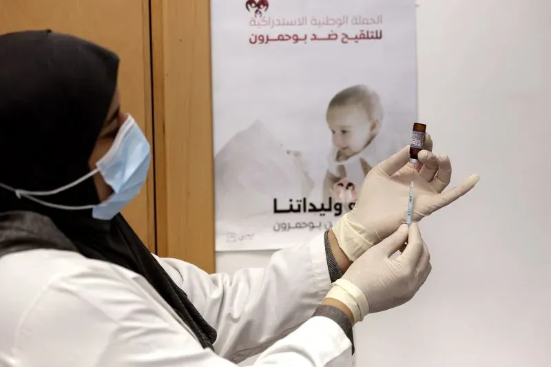 مقدمة لموظفة صحية ترتدي قناعًا وتستعد لتطعيم الأطفال ضد الحصبة، مع وجود ملصق توعوي خلفها حول أهمية التطعيم.