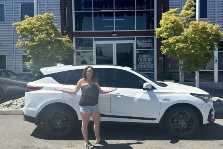 امرأة تقف بجانب سيارة أكيورا RDX بيضاء في ساحة عرض سيارات، تعبر عن فرحتها بعد شراء سيارة جديدة وسط ارتفاع أسعار السيارات.