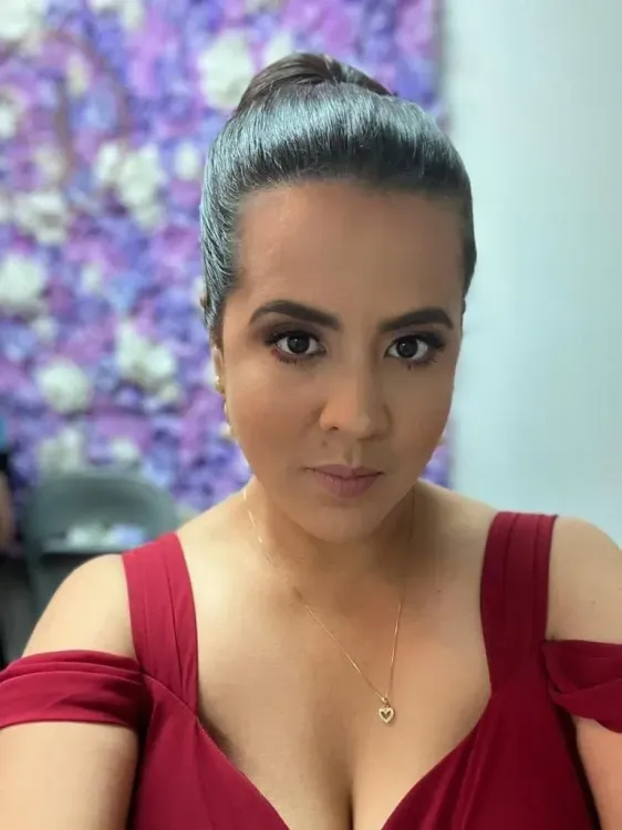 إستيفاني رودريغيز، صحفية كولومبية، تظهر في صورة سيلفي، ترتدي فستانًا أحمر، مع خلفية مزهرة. تغطي اعتقالات المهاجرين.