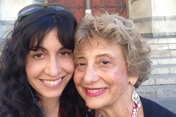 صورة لامرأتين مبتسمتين، الأم وابنتها، تتشاركان لحظة حميمية. تعكس الصورة حبًا عميقًا وعلاقة قوية بينهما.