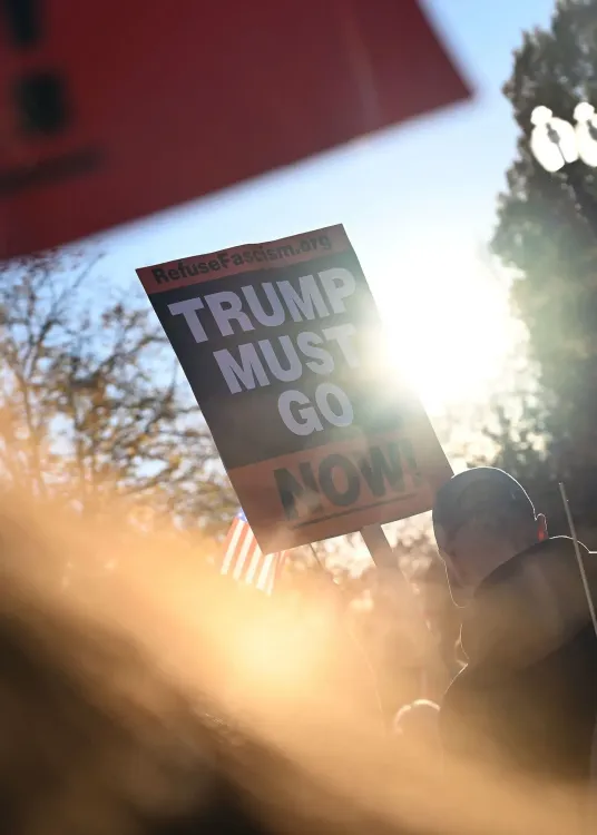 لافتة مكتوب عليها "ترامب يجب أن يرحل الآن!" في تجمع احتجاجي، مع أشعة الشمس تتسلل من الخلف، مما يبرز الأجواء المتوترة.