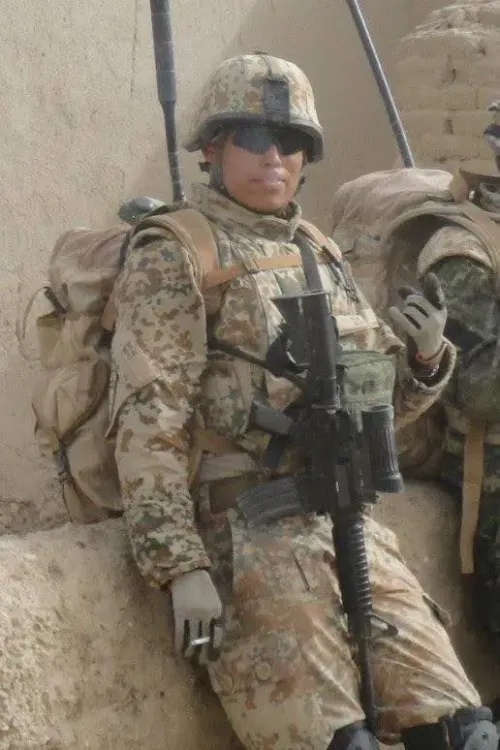 جندي يرتدي زيًا عسكريًا في منطقة صحراوية، يحمل سلاحًا ويظهر تعبيرًا غير رسمي، مما يعكس تجربة الخدمة العسكرية في بيئة صعبة.