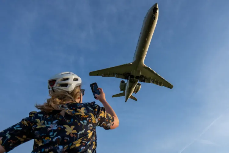 امرأة ترتدي خوذة تلتقط صورة لطائرة تحلق في السماء، مما يعكس تأثير ارتفاع أسعار وقود الطائرات على السفر الجوي.