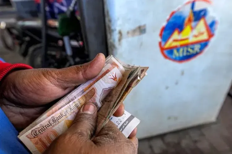 رجل يحمل نقودًا من فئات مختلفة أمام محطة وقود، مما يعكس تأثير ارتفاع أسعار الطاقة على الاقتصاد في الدول النامية.
