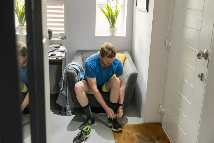 رجل يجلس على أريكة في منزله، يرتدي ملابس رياضية ويقوم بربط حذائه استعدادًا لممارسة الرياضة، مما يعكس أهمية النشاط البدني.