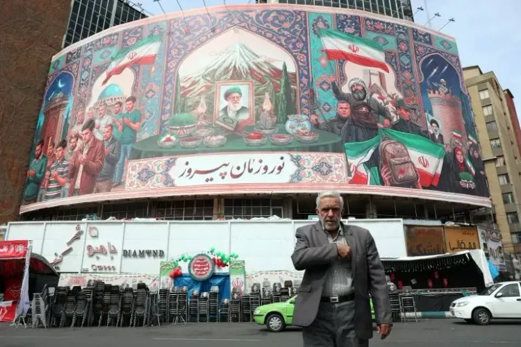 رجل مسن يسير أمام جدارية كبيرة في طهران تُظهر مشاهد ثقافية إيرانية، مع العلم الإيراني في الخلفية، تعكس التوترات الإقليمية.