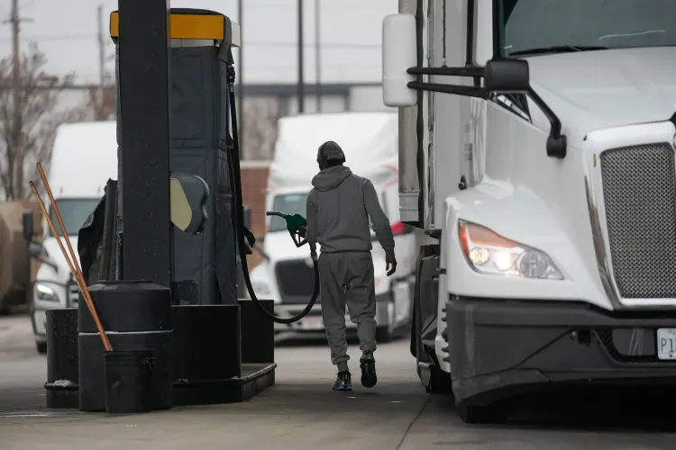 رجل يرتدي ملابس رمادية يتوجه نحو مضخة وقود في محطة، مع شاحنة كبيرة في الخلفية، مما يعكس تأثير ارتفاع أسعار الوقود على الاقتصاد الأمريكي.