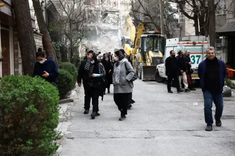 مشهد لشارع في طهران بعد غارة، حيث يسير الناس بحذر وسط الدمار، مع وجود معدات إنقاذ في الخلفية.