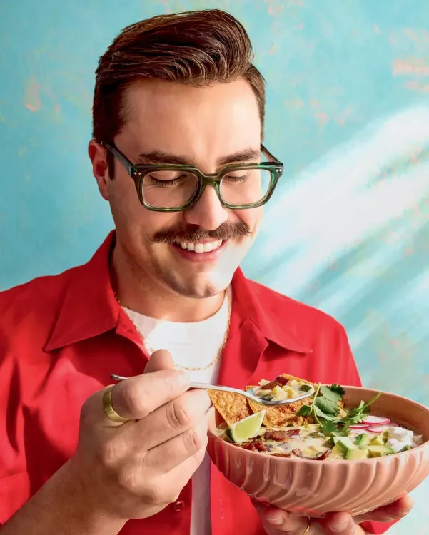 طاهٍ يحمل وعاءً من الطعام الشهي، مبتسمًا، مع خلفية ملونة تعكس أجواء الطهي الإبداعي والمكونات الموسمية.