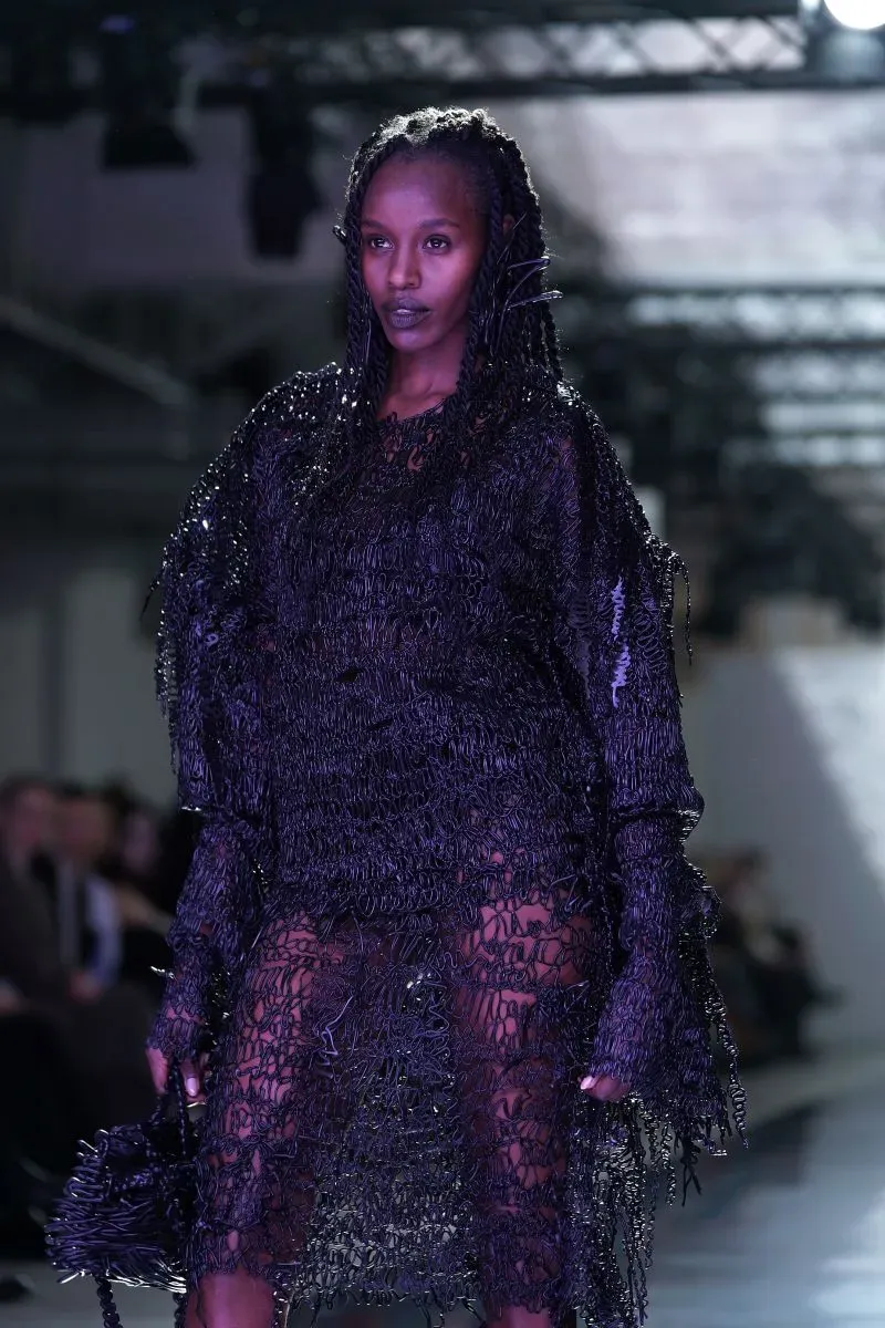 عارضة أزياء ترتدي فستانًا أسود بلا أكمام مصنوع من اللاتكس المطبوعة بتقنية ثلاثية الأبعاد، مع تفاصيل معقدة تشبه الدانتيل.