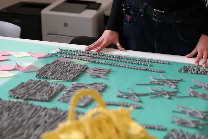 يد تظهر يد مصممة الأزياء كاسيا كوتشارسكا وهي تتعامل مع قطع من اللاتكس الملون على طاولة عمل، مع أدوات خياطة وخلفية مكتبية.