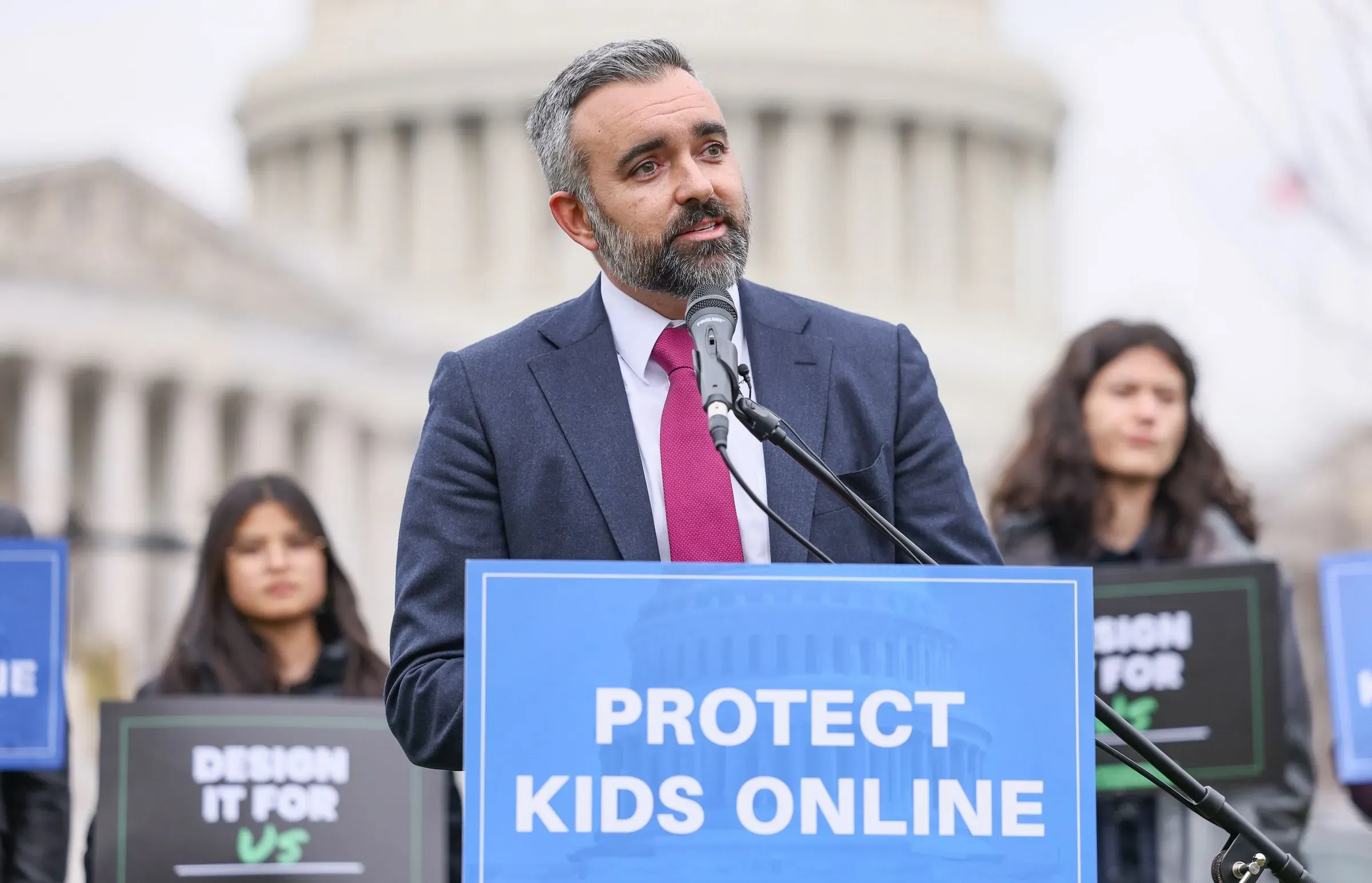 رجل يتحدث في مؤتمر صحفي أمام مبنى حكومي، يحمل لافتة تطالب بحماية الأطفال عبر الإنترنت، مع وجود متظاهرين خلفه.