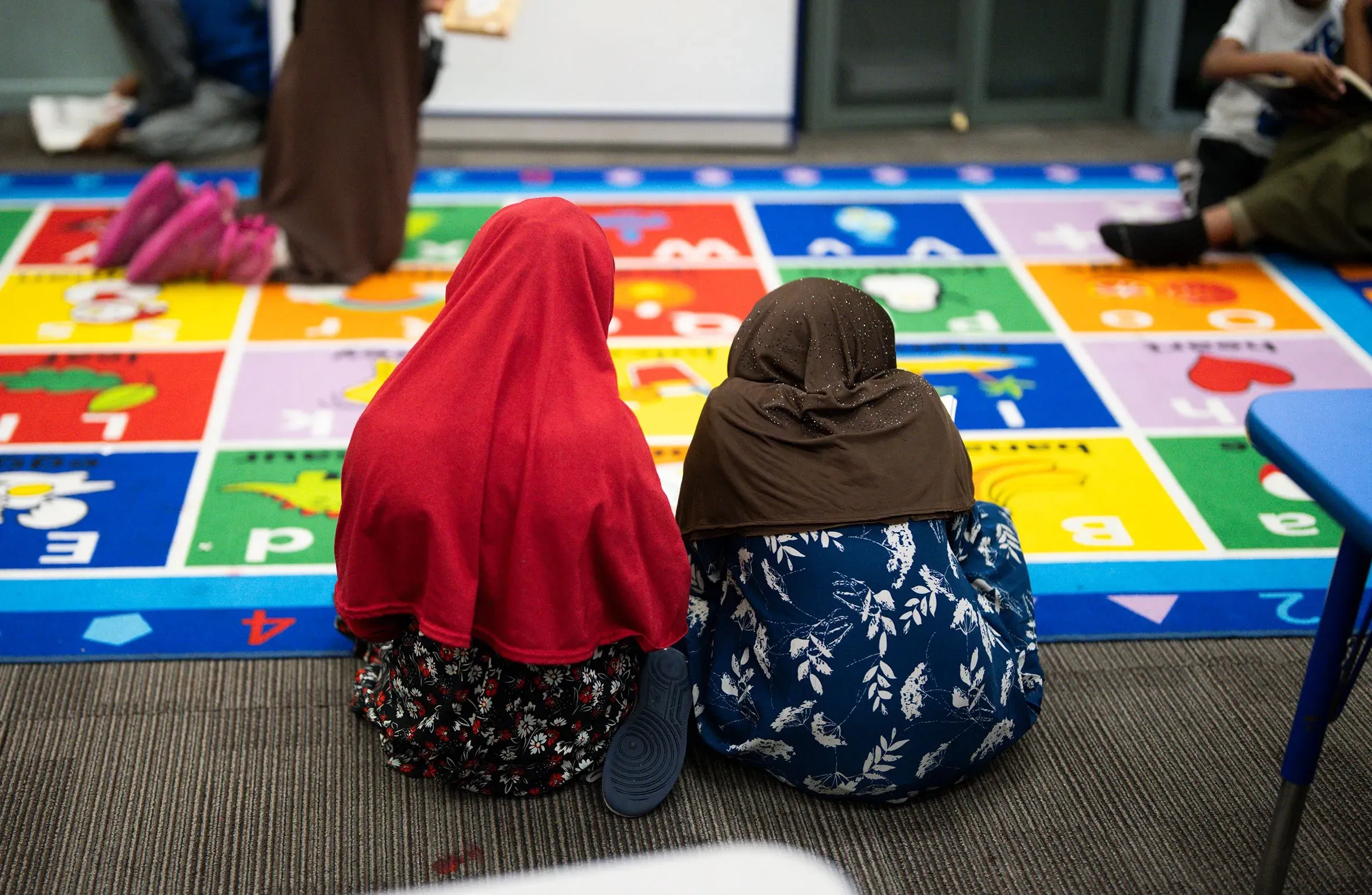 طفلتان ترتديان الحجاب تجلسان معًا على سجادة ملونة في مركز رعاية أطفال، مع وجود أطفال آخرين في الخلفية.