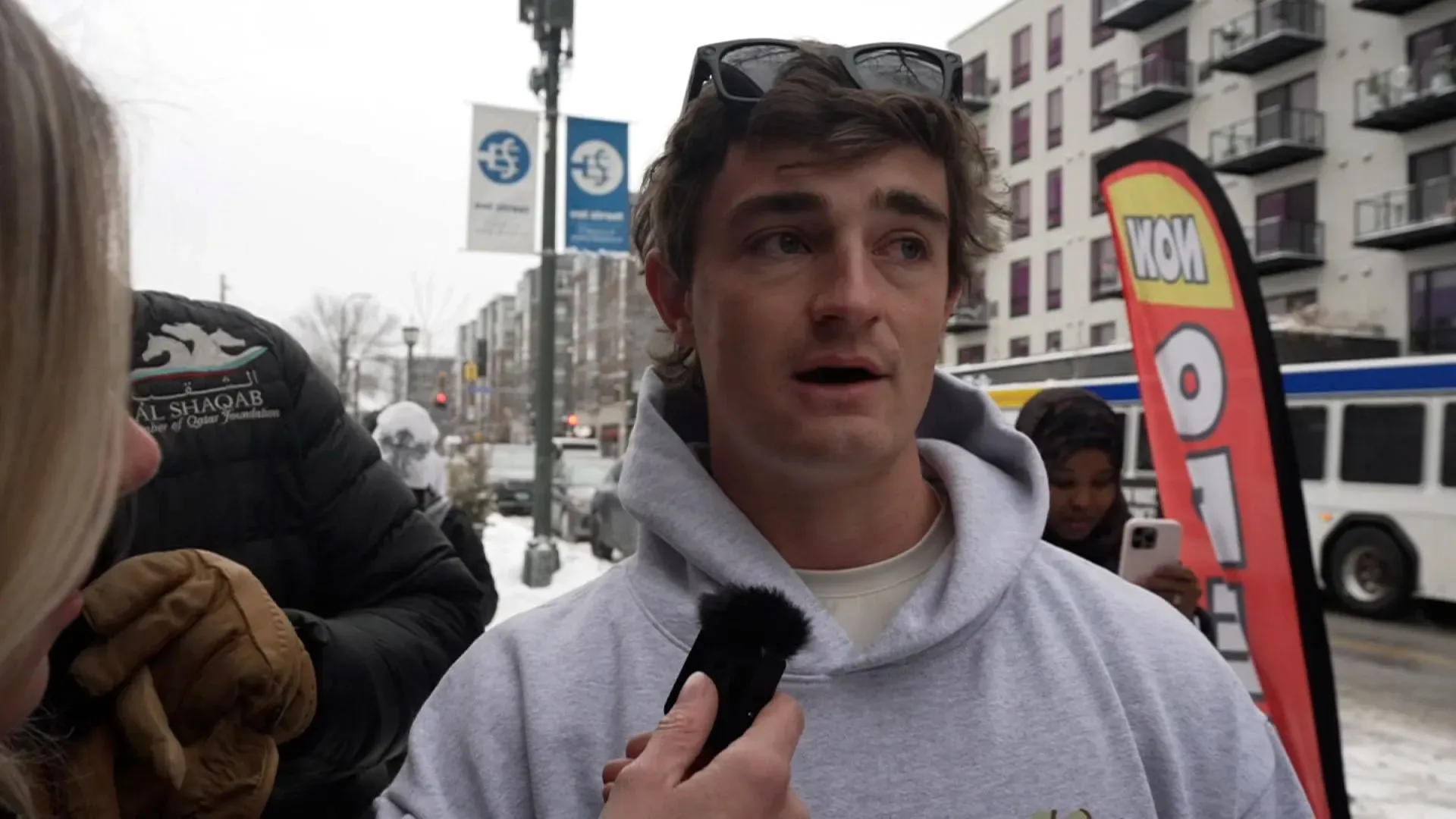 شخص يتحدث في الشارع خلال طقس ثلجي، مع لافتة تشير إلى حدث محلي، بينما تظهر مبانٍ خلفه.