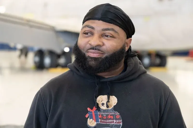 رجل مبتسم يرتدي سترة سوداء تحمل صورة دب، يقف في مكان مفتوح مع طائرات خلفه، يعكس روح الأمل والتعافي بعد مأساة.