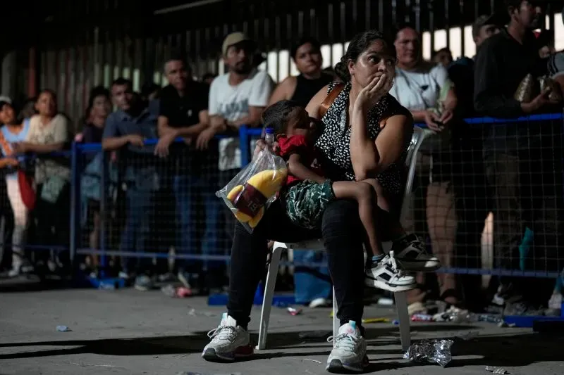 امرأة تحمل طفلاً يجلسان على كرسي في منطقة مزدحمة، مع طوابير طويلة في الخلفية، تعبير يرسم قلق النازحين من العنف في كولومبيا.