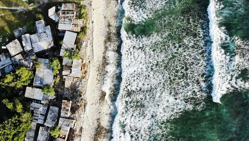 صورة جوية تظهر منازل مدمرة على شاطئ جزيرة توفالو، مع أمواج البحر تتلاطم على الرمال، تعكس تأثير تغير المناخ وارتفاع منسوب المياه.