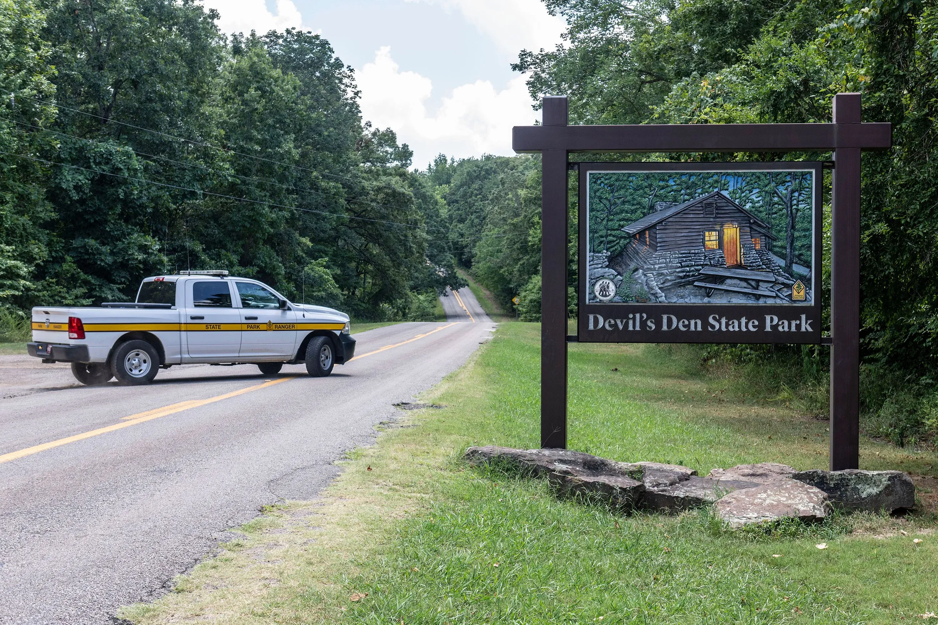لافتة تشير إلى متنزه Devil's Den State Park، مع شاحنة تابعة للشرطة في الطريق، محاطة بالأشجار الكثيفة.