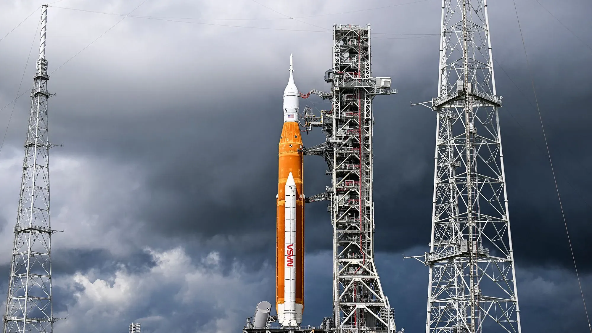 صاروخ SLS التابع لناسا، ذو اللون البرتقالي، واقفًا على منصة الإطلاق تحت سماء غائمة، يمثل جزءًا أساسيًا من برنامج أرتميس لاستكشاف القمر.