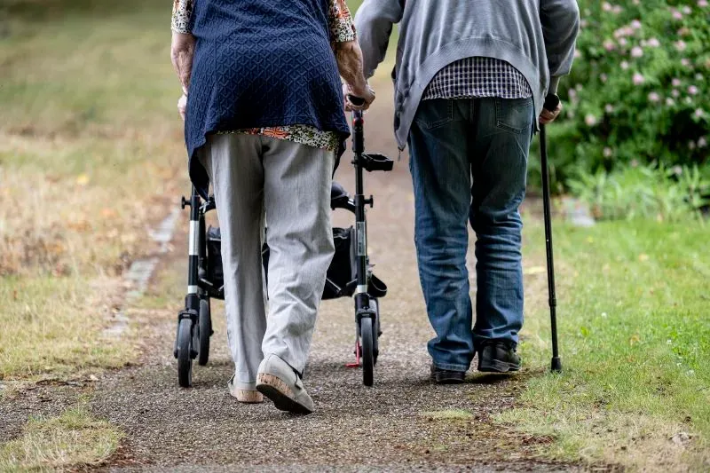شخصان مسنّان يسيران معًا على ممر، أحدهما يستخدم عكازًا والآخر يدفع عربة مساعدة، مما يعكس التحديات المالية والاجتماعية في أوروبا.