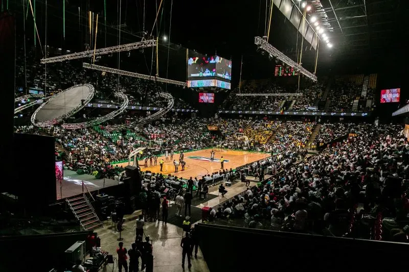 مدرج مزدحم في صالة رياضية خلال مباراة كرة سلة، مع لاعبين يتنافسون في الملعب وشاشات عرض كبيرة تعرض المباراة.