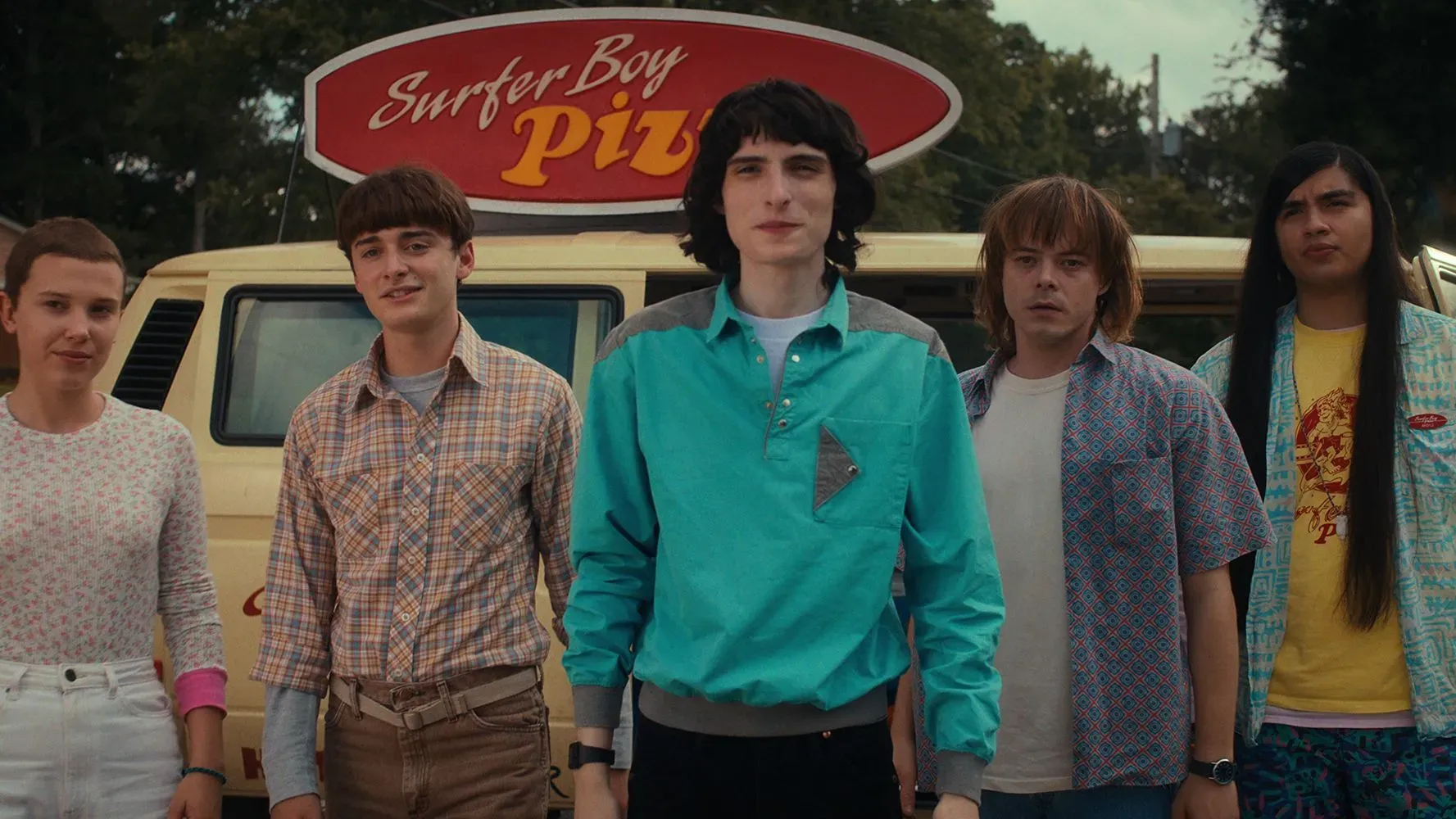 مجموعة من الشخصيات الرئيسية من مسلسل "Stranger Things" يقفون أمام شاحنة بيتزا تحمل شعار "Surfer Boy Pizza"، مع خلفية تعكس أجواء الثمانينات.