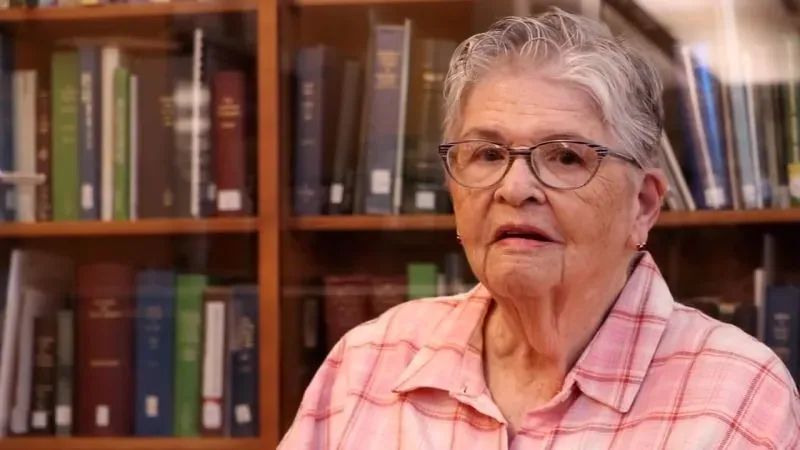 امرأة مسنّة تجلس أمام رفوف مكتبة مليئة بالكتب، تعبر عن فرحتها بعد إعادة كتاب قديم يعود لعام 1926.