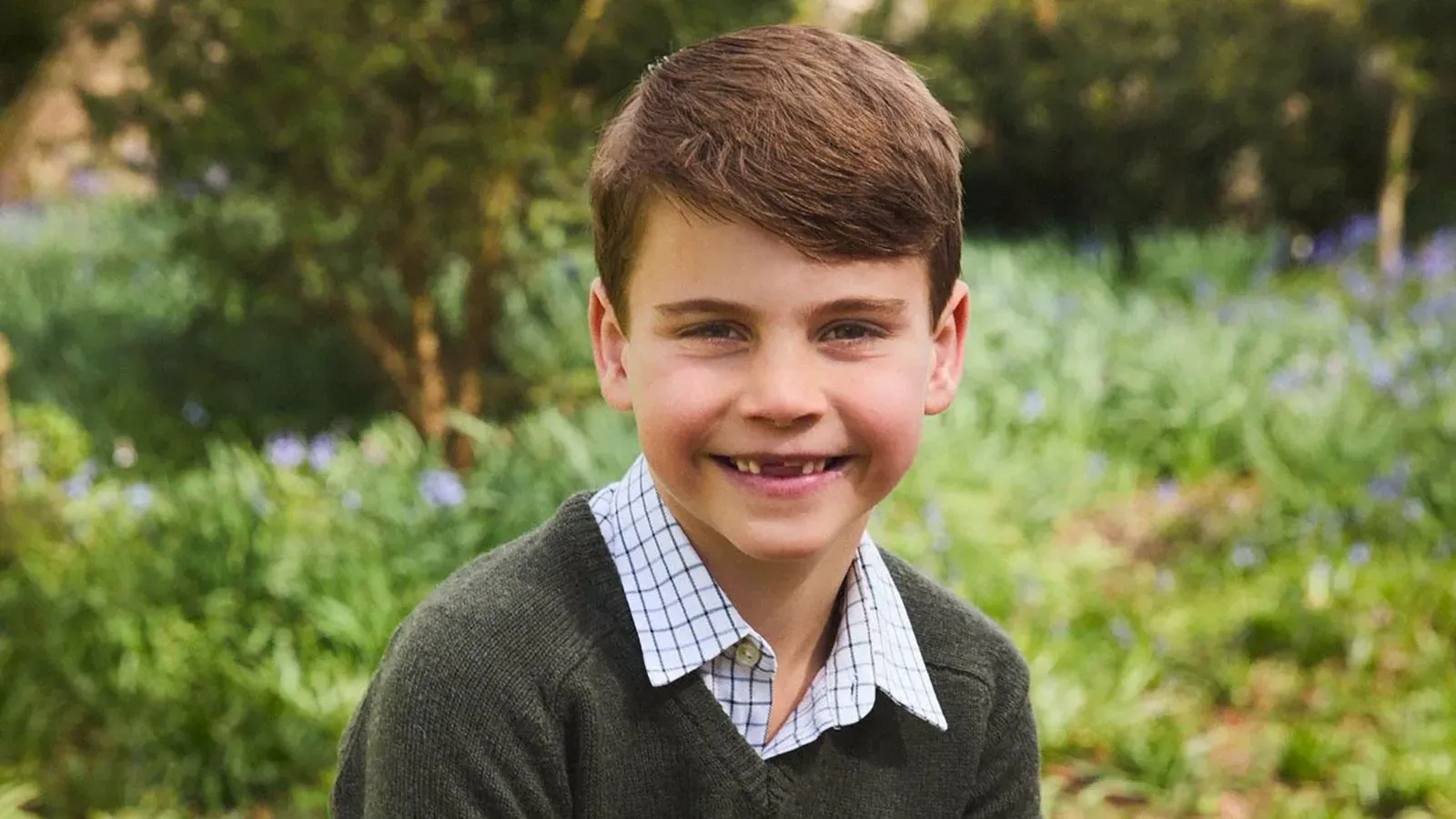 الأمير لويس، في السابعة من عمره، مبتسمًا بجذع شجرة، يرتدي قميصًا بياقة وسترة خضراء، مع فقدان أسنان أمامية.
