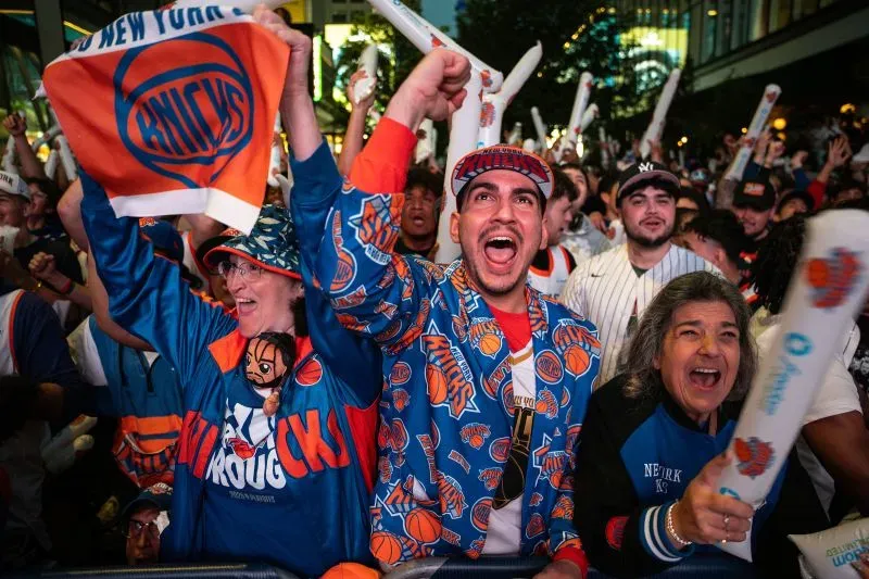 احتفالات حماسية لمشجعي نيويورك نيكس في الشوارع بعد فوز الفريق على بوسطن سيلتيكس، مع شعارات وأعلام تعبر عن الفخر والفرحة.