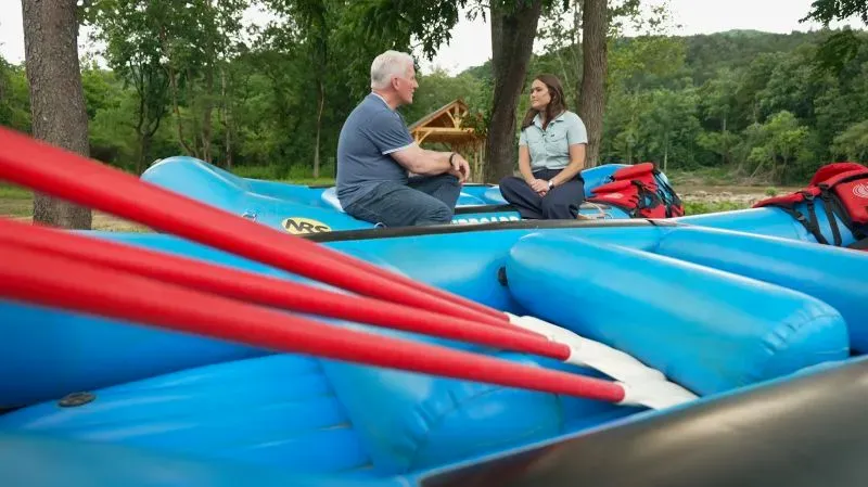 مقابلة بين كوري هامبتون وشخص آخر على قوارب زرقاء قرب نهر فرينش برود، مع أشجار خضراء في الخلفية، تعكس جهود التعافي بعد إعصار هيلين.