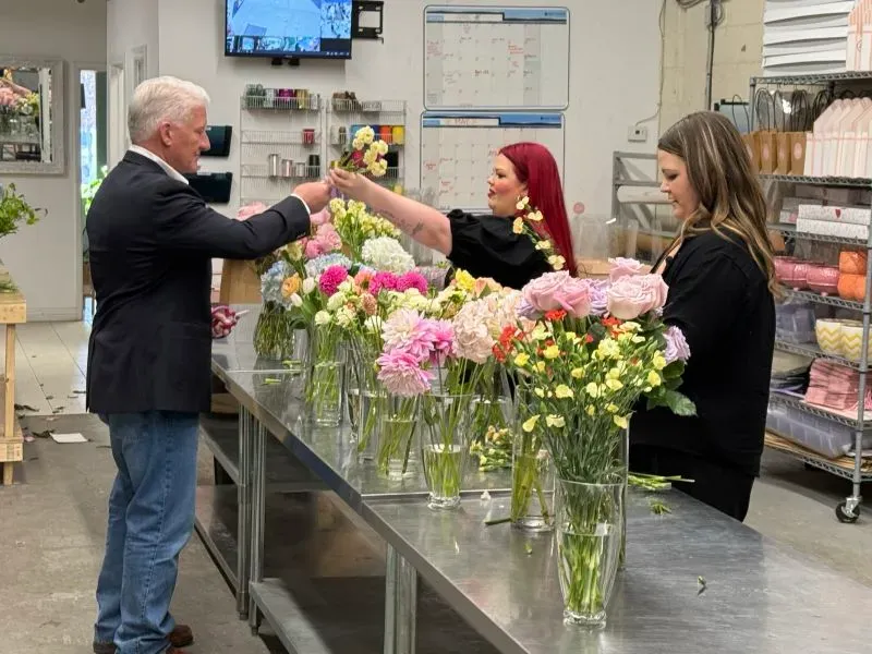 شخصية تتفاعل مع عاملتين في محل زهور، حيث ينسقون أزهار ملونة مثل الداليا والقرنفل، وسط أجواء عمل نشطة.