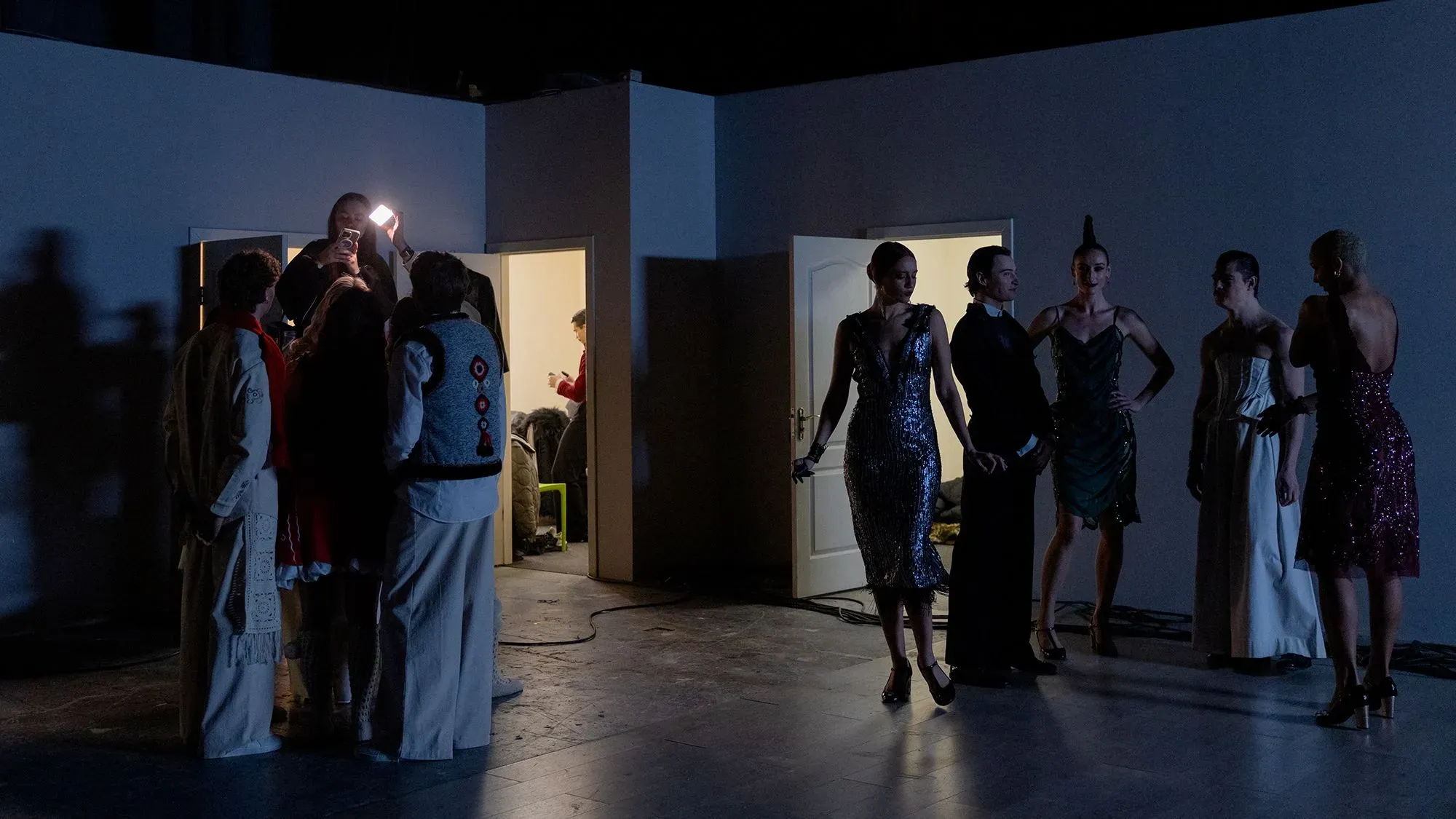 مجموعة من الأشخاص يرتدون ملابس أنيقة في كواليس عرض أزياء، مع إضاءة خافتة وظلال واضحة، مما يعكس أجواء التحضير للعرض.