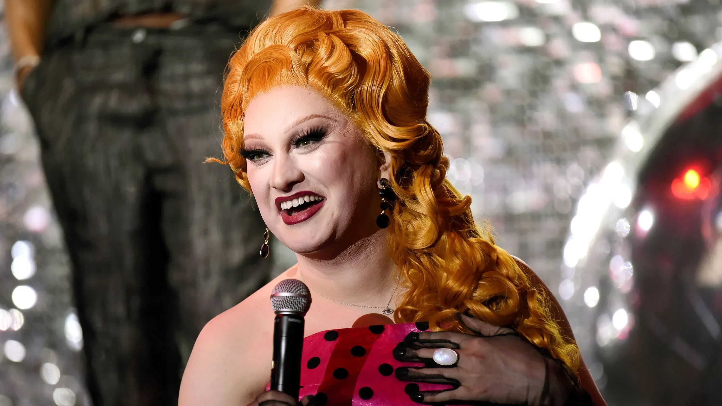 جينكس مونسون، نجمة "سباق السحرة روبول"، تبتسم أثناء حديثها في عرض مسرحي، مع تسريحة شعر برتقالية وملابس ملونة.