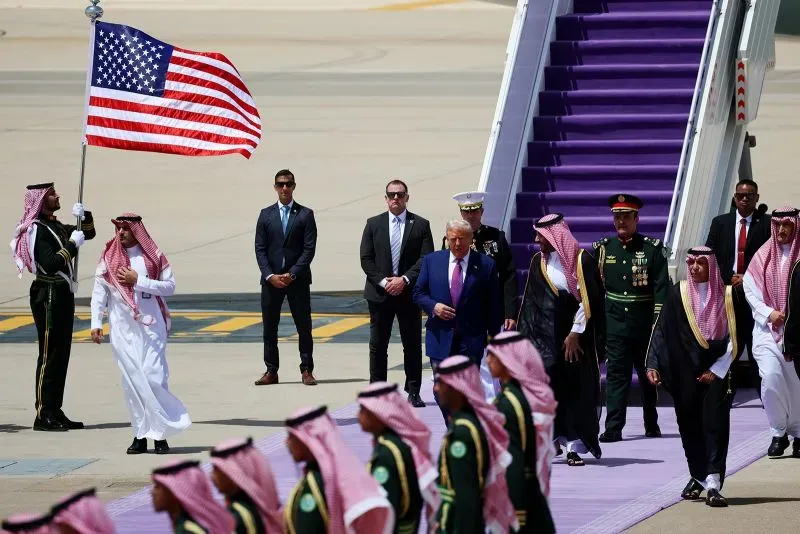 ترامب يستقبل في السعودية مع وفد من رجال الأعمال، بينما يحمل أحدهم علم الولايات المتحدة، وسط مراسم رسمية.