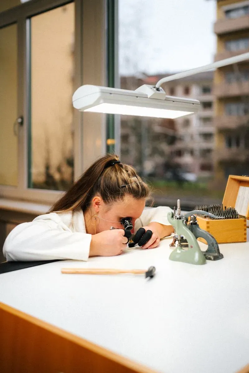 شابة ترتدي معطفًا أبيض، تركز على تفاصيل دقيقة باستخدام مجهر، في ورشة لصناعة الساعات، تعكس اهتمام الجيل الجديد بهذه الحرفة التقليدية.