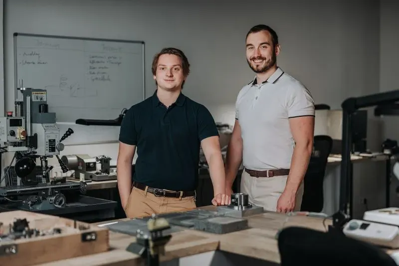 شابان مبتسمان يقفان في ورشة لصناعة الساعات، محاطين بأدوات وآلات دقيقة، تعكس اهتمام الجيل الجديد بالحرفة التقليدية.