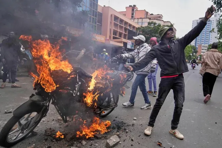 شخص يحمل هاتفه وسط احتجاجات في نيروبي، بينما تشتعل دراجة نارية بالقرب منه، مما يعكس التوترات المتزايدة ضد عنف الشرطة.
