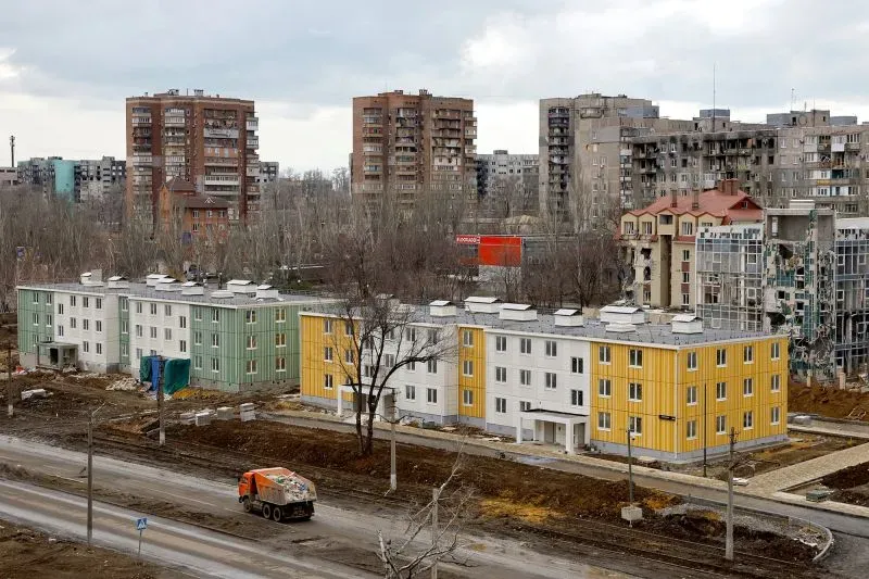 مشهد لعدة مبانٍ سكنية جديدة بألوان مختلفة في ماريوبول، مع خلفية لمباني متضررة، يعكس جهود إعادة الإعمار تحت الاحتلال الروسي.