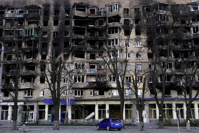 مبنى سكني متضرر في ماريوبول، يظهر آثار الدمار نتيجة الحرب، مع نوافذ محطمة وأشجار جرداء في الواجهة.